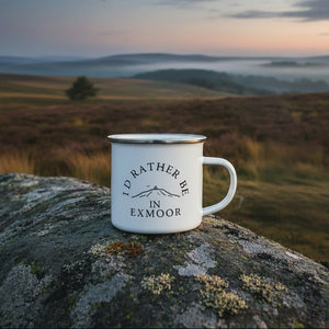 A white enamel-style mug with a silver rim and black text reading 'I'D RATHER BE IN EXMOOR' with a simple mountain illustration, placed on a plaid blanket in neutral and red tones.