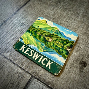 A square coaster featuring a vintage-style illustration of Keswick, with green hills, blue water, and a dark green banner reading 'KESWICK' in white letters, placed on a weathered gray wooden surface.