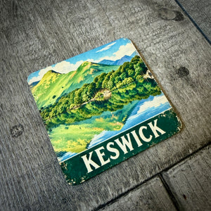 A square coaster featuring a vintage-style illustration of Keswick, with green hills, blue water, and a dark green band at the bottom reading 'KESWICK' in white letters, placed on a weathered gray wooden surface.
