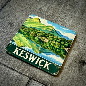 A square coaster featuring a vintage-style illustration of Keswick, Scotland, with green hills, blue water, and the word 'KESWICK' in white on a dark green banner, placed on a weathered gray wooden surface.
