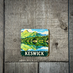 A square coaster featuring a vintage-style illustration of Keswick, UK, with green hills, a blue lake reflection, and the text 'KESWICK' at the bottom, displayed on a rustic gray wooden background.