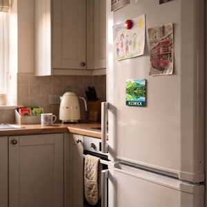 A white refrigerator in a real kitchen setting, decorated with several colorful magnets and children's drawings. The refrigerator door has a green 'KESWICK' magnet, and the kitchen features cream-colored cabinets, a tiled backsplash, and a wooden countertop.