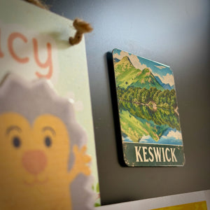 A small rectangular Keswick landscape magnet with a mountain lake scene and the word 'KESWICK' at the bottom, photographed against a dark gray surface. Part of a colorful hanging card with a cartoon face is blurred in the foreground.